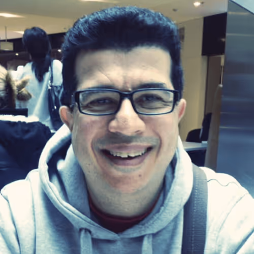 Smiling man wearing glasses and a grey hoodie in a public indoor space.