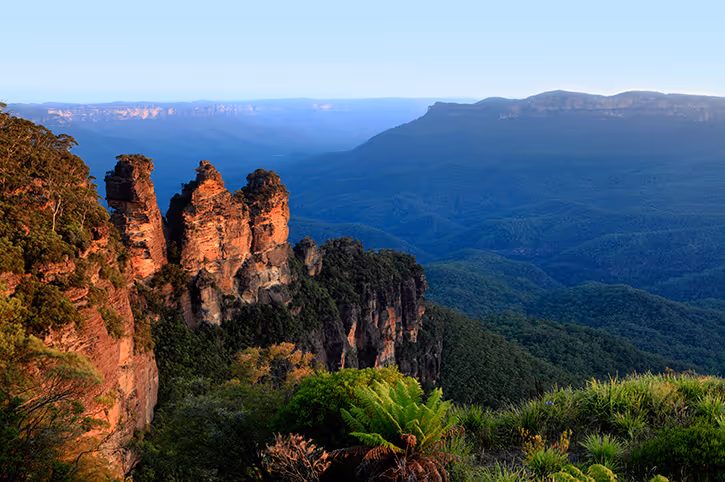 The Three Sisters rock formation at Blue Mountains with surrounding lush green forest under a clear blue sky.