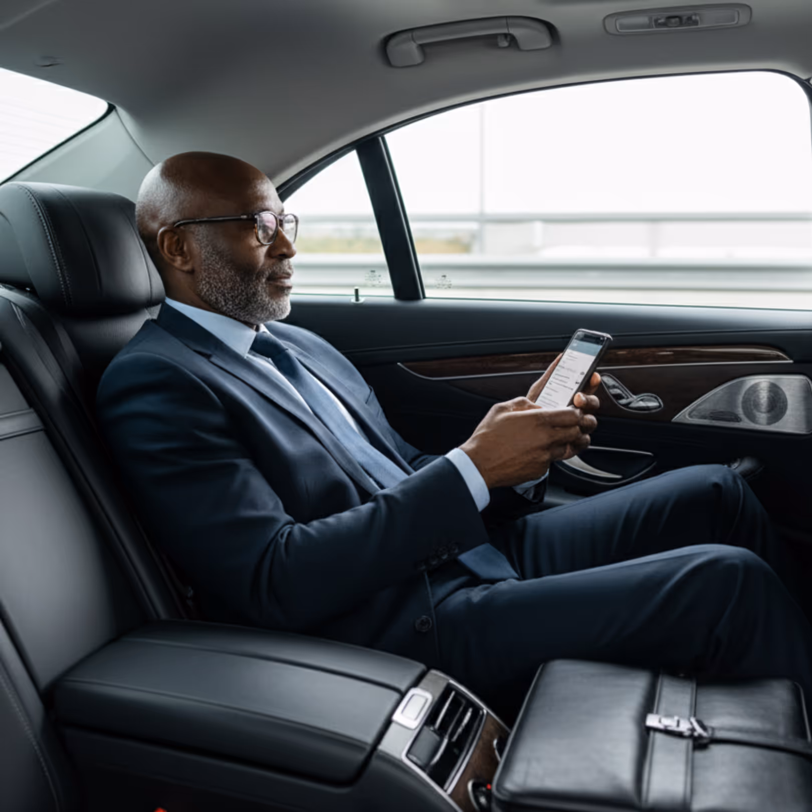 A senior executive sits comfortably in the back seat of a luxury car, using his smartphone to check emails during his professional airport transfer. The vehicle's clean interior and tinted windows convey privacy and comfort.