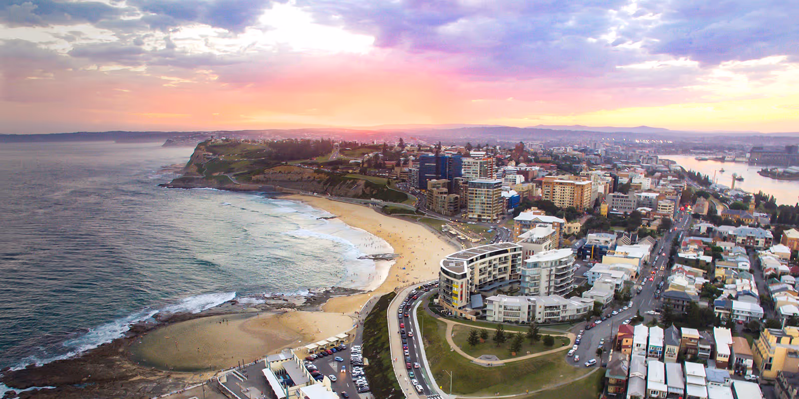 Aerial view of a coastal city at sunset featuring a curved sandy beach, residential buildings, and a road along the shoreline.