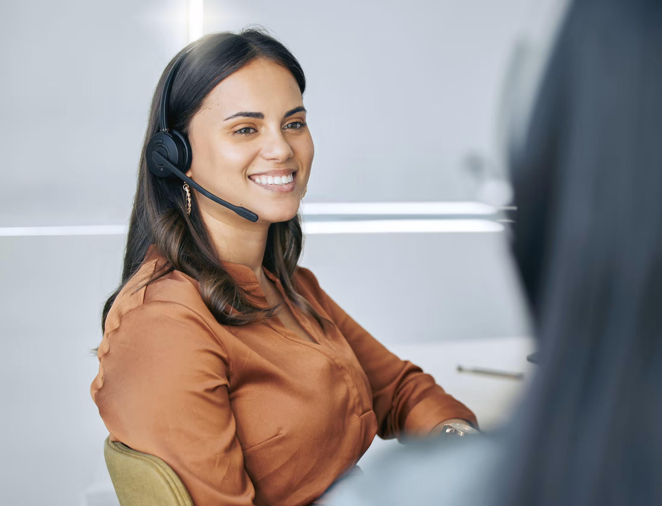 Smiling woman wearing a headset, seated and engaged in conversation in an office setting.