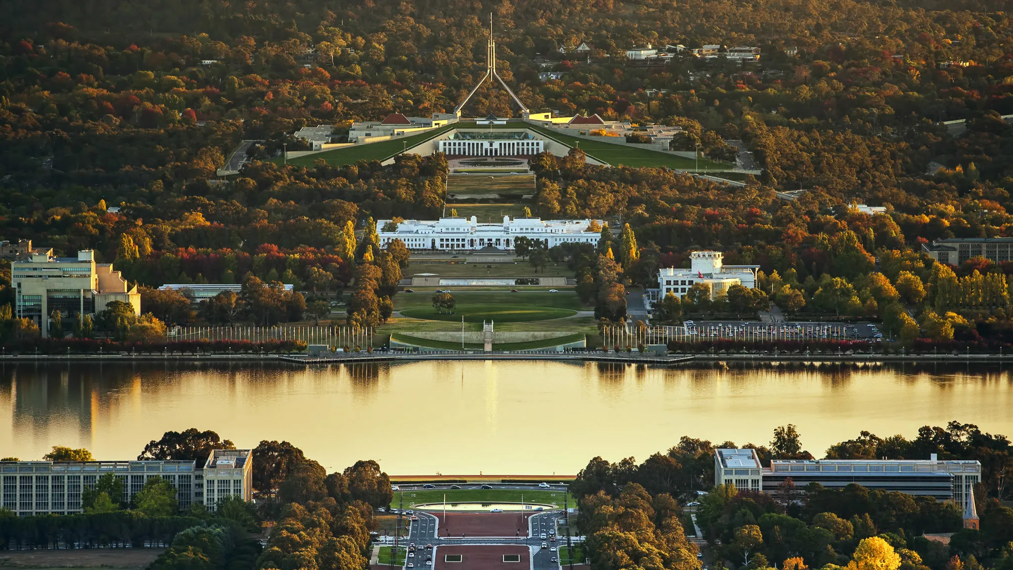 Aerial view of Canberra showing the Parliament House atop a hill, the surrounding greenery, and a reflective lake in the foreground.