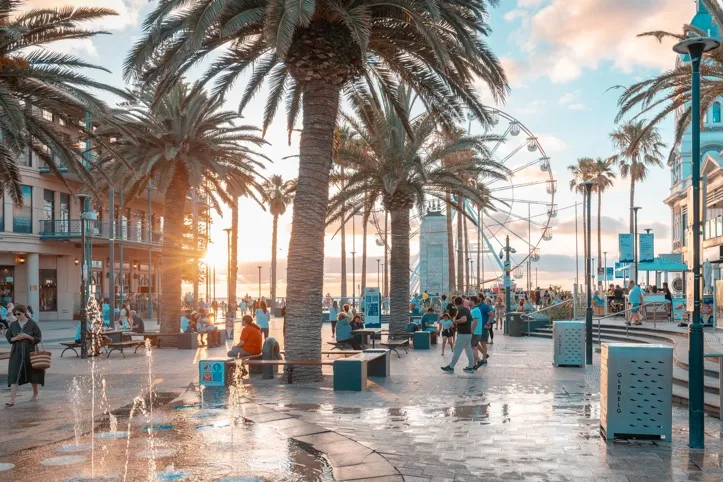 Crowded oceanfront promenade with palm trees, people walking, a fountain, and a Ferris wheel at sunset.