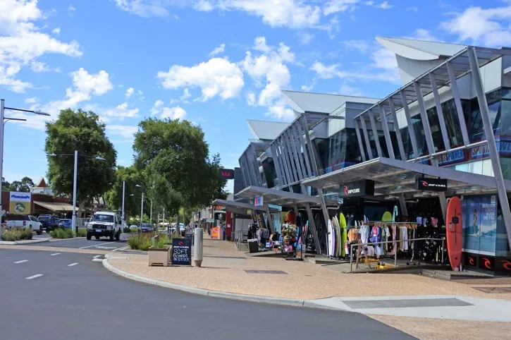 Street view of a shopping area featuring a surf shop with surfboards and clothing displayed outside under a modern glass and metal awning, with trees and a blue sky in the background.