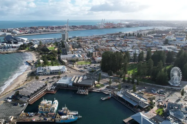 Aerial view of Fremantle Harbour area with docks, buildings, a Ferris wheel, and the ocean under a partly cloudy sky.