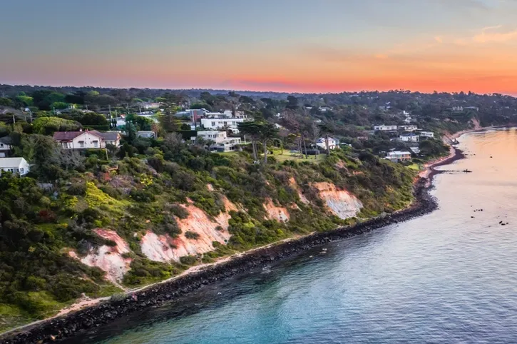 Coastal cliffside with residential houses overlooking calm ocean waters at sunset.