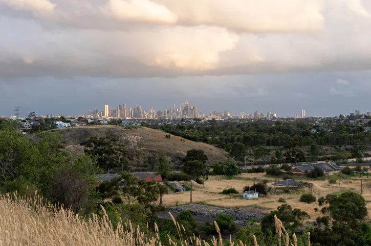 View of a grassy hill and trees in the foreground with a city skyline under a cloudy sky in the distance.