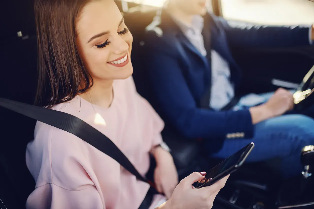 Smiling passenger checking her phone in the back of a chauffeur-driven car on the way to the airport