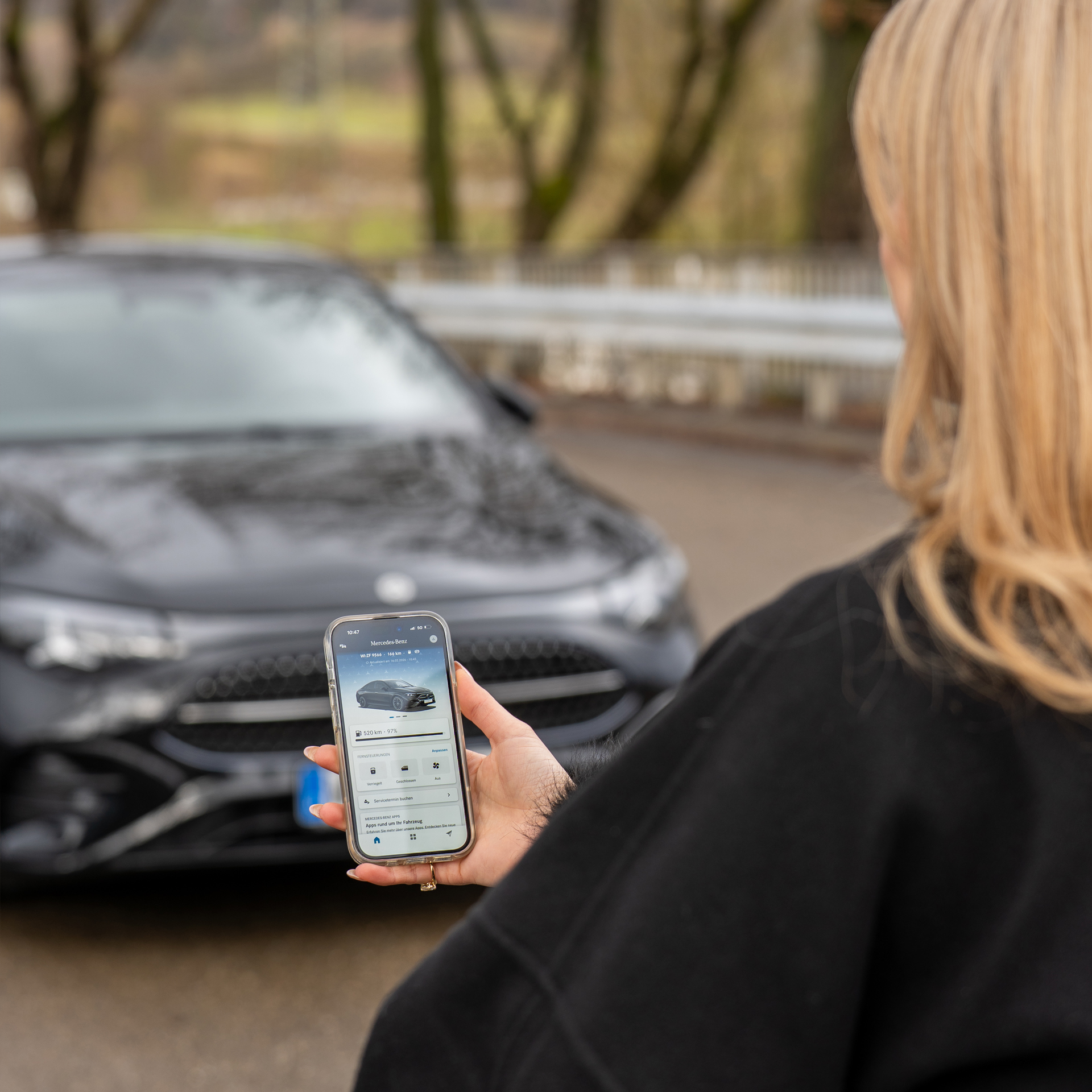 Bildmotiv zum Thema "Digitale Extras". Man sieht einen Schwarzen Mercedes-Benz Pkw im Hintergrund und vorn rechts im Bild eine Frau von hinten mit langen blonden Haaren, einer Schwarzen Jacke und einem Smartphone in der Hand.
