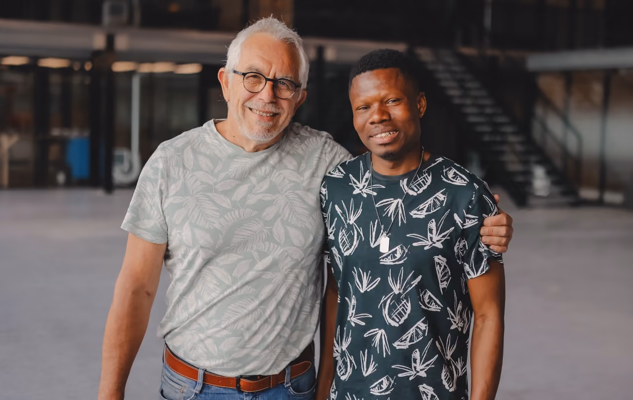 Twee mensen glimlachen naar de fotograaf, de mentor: een 60-jarige man met kort, wit haar en een bril sloeg zijn arm om de schouders van de mentee, een glimlachende jonge zwarte man.