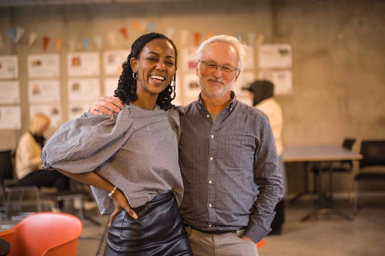 Une jeune femme noire pose avec un grand sourire aux côtés de son mentor un homme aux cheveux blancs avec des lunettes