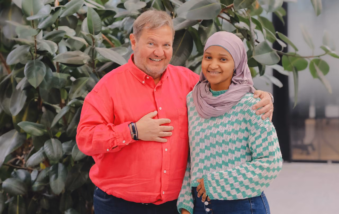 Un homme d'une cinquantaine d'années pose en souriant avec sa mentee, une jeune femme avec un voile mauve qui sourit aussi