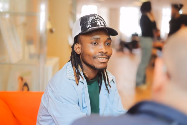 Un jeune homme noir avec un casquette et des locks sourit à une personne hors cadre