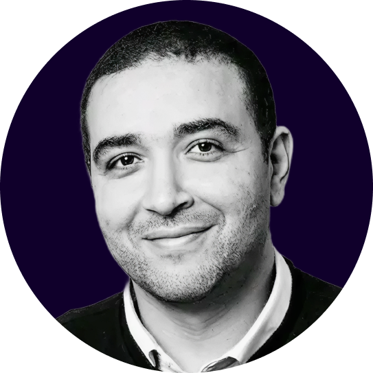 Portrait en noir et blanc d'un homme souriant avec un léger bouc, portant une chemise blanche et un pull sombre.