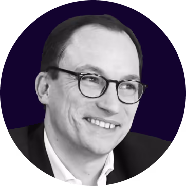 Portrait d'un homme souriant portant des lunettes et un costume noir sur fond sombre.