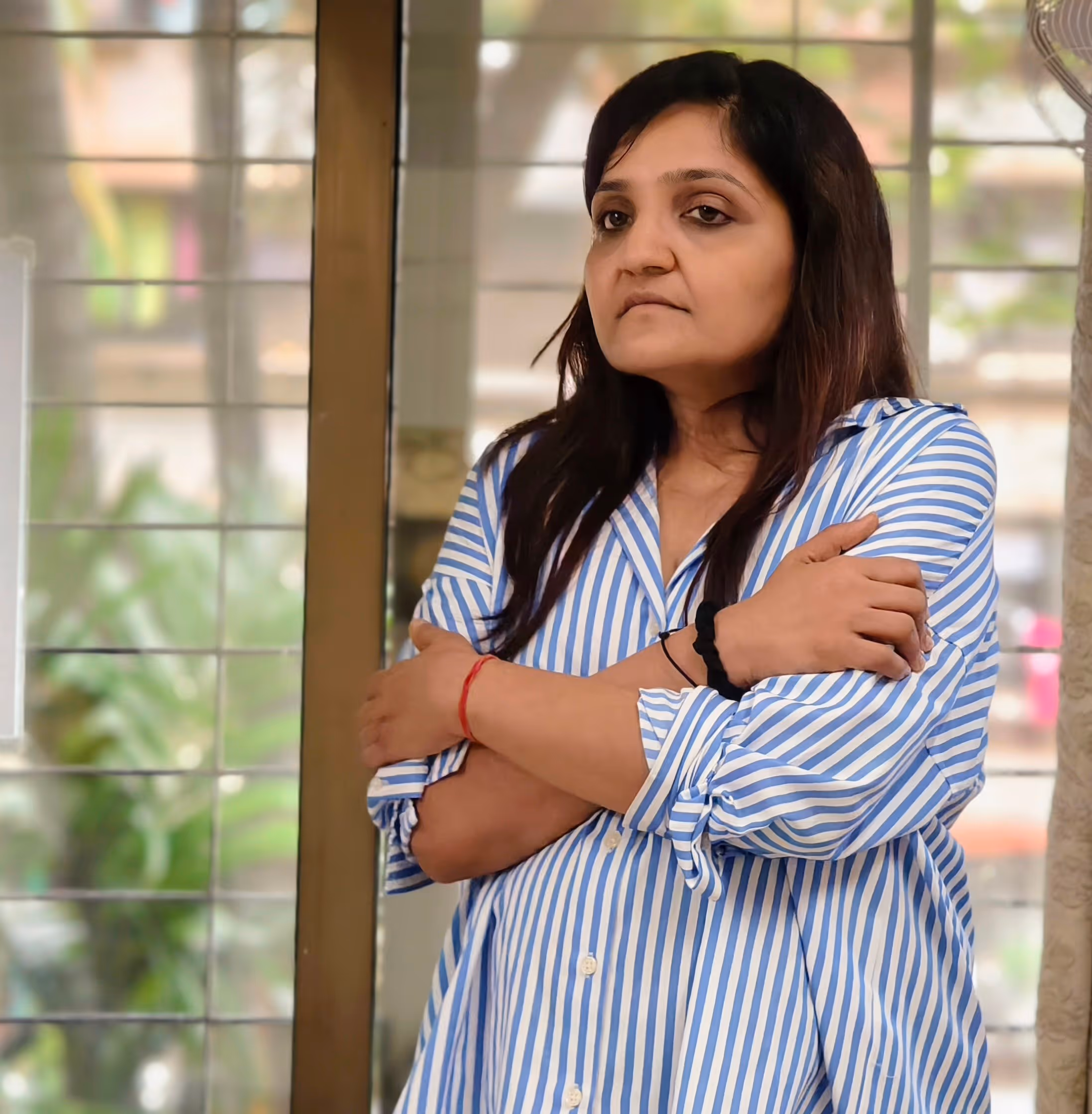Woman in blue and white striped shirt crossing her arms, standing indoors near a window, during NLP Training Course.