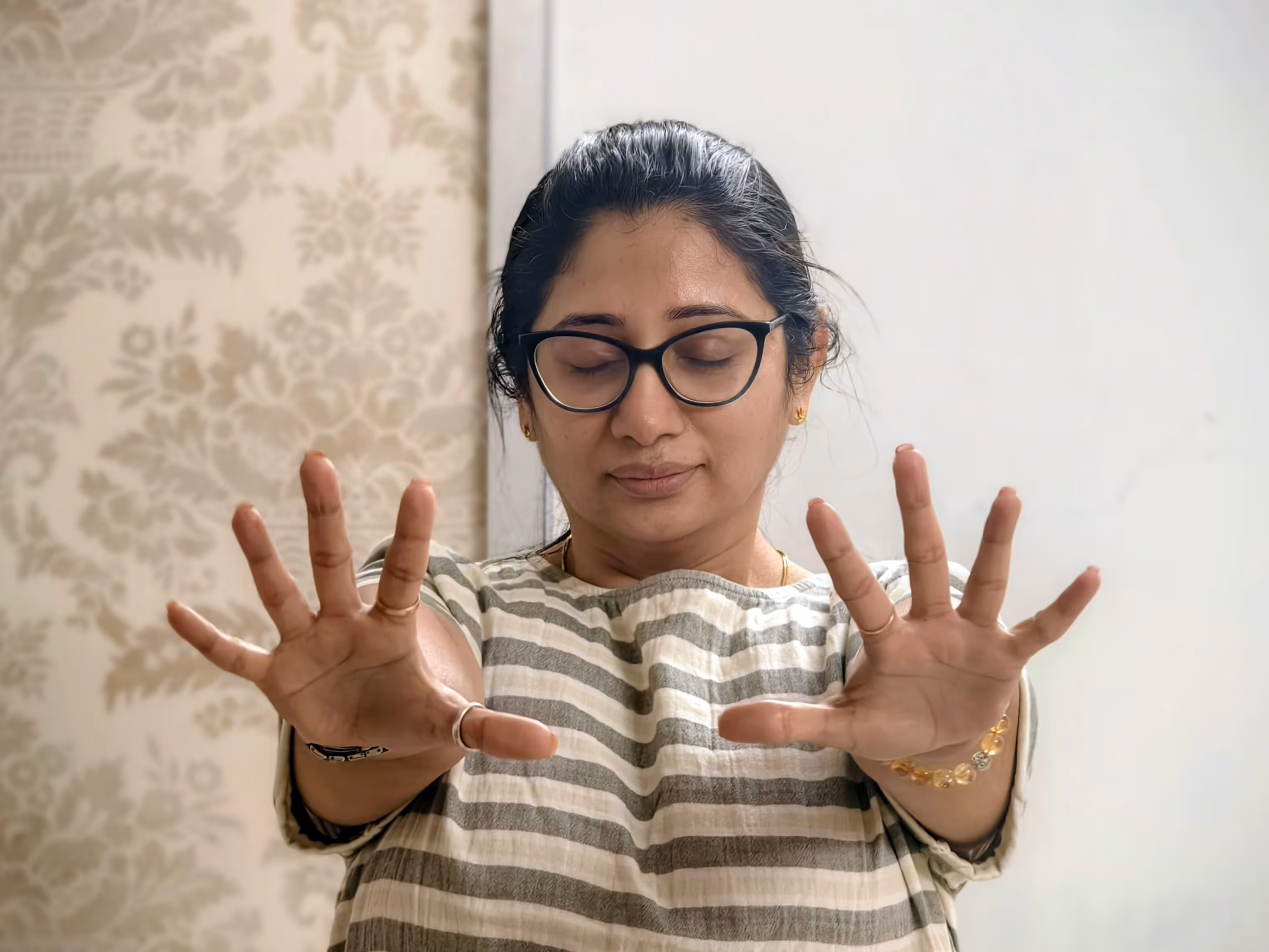 Woman with glasses and closed eyes stretches her hands forward wearing striped shirt, during Life Coach certification course in India.