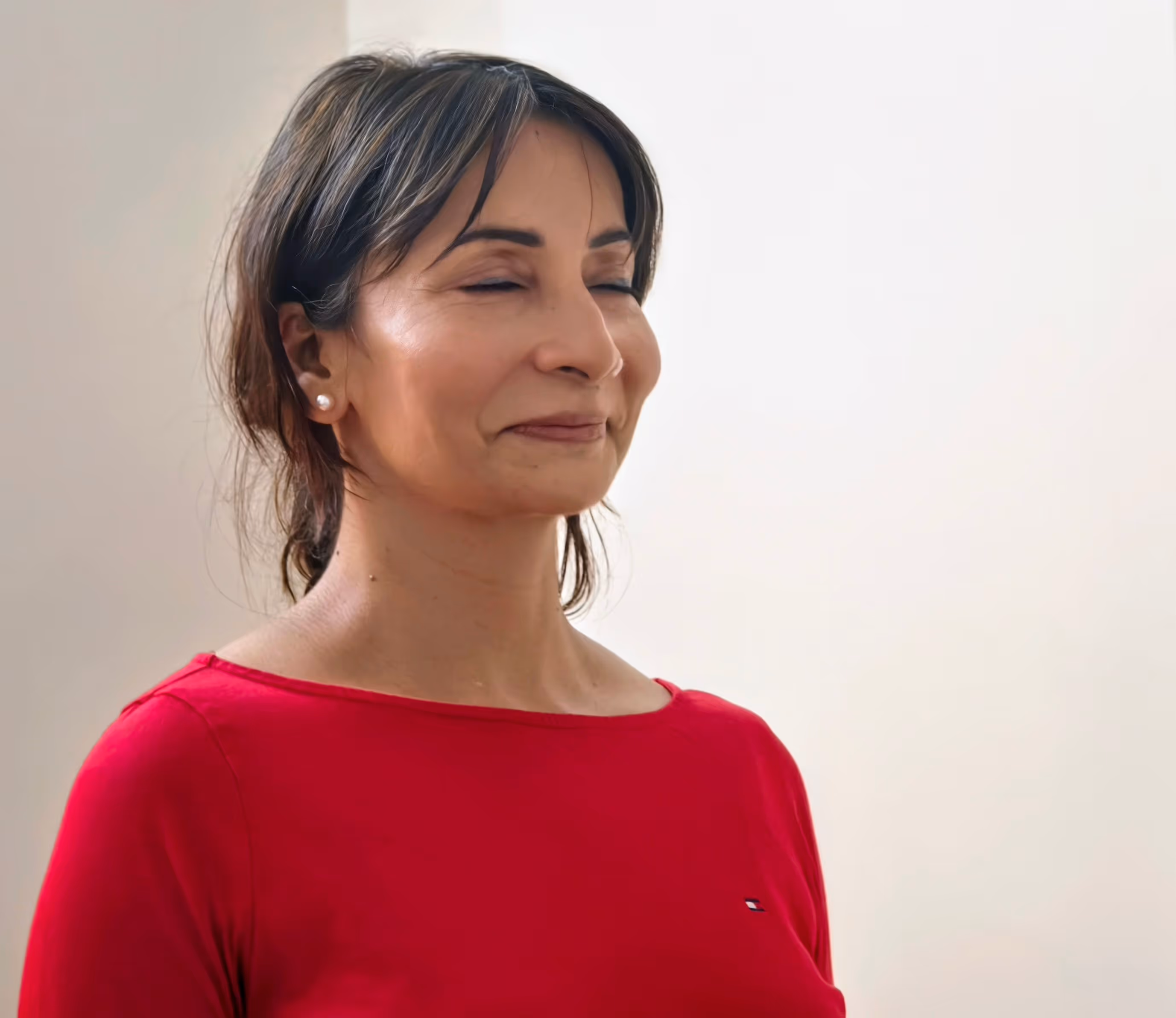 Smiling woman with closed eyes wearing red top and pearl earrings against plain background, during Abhay's NLP course.