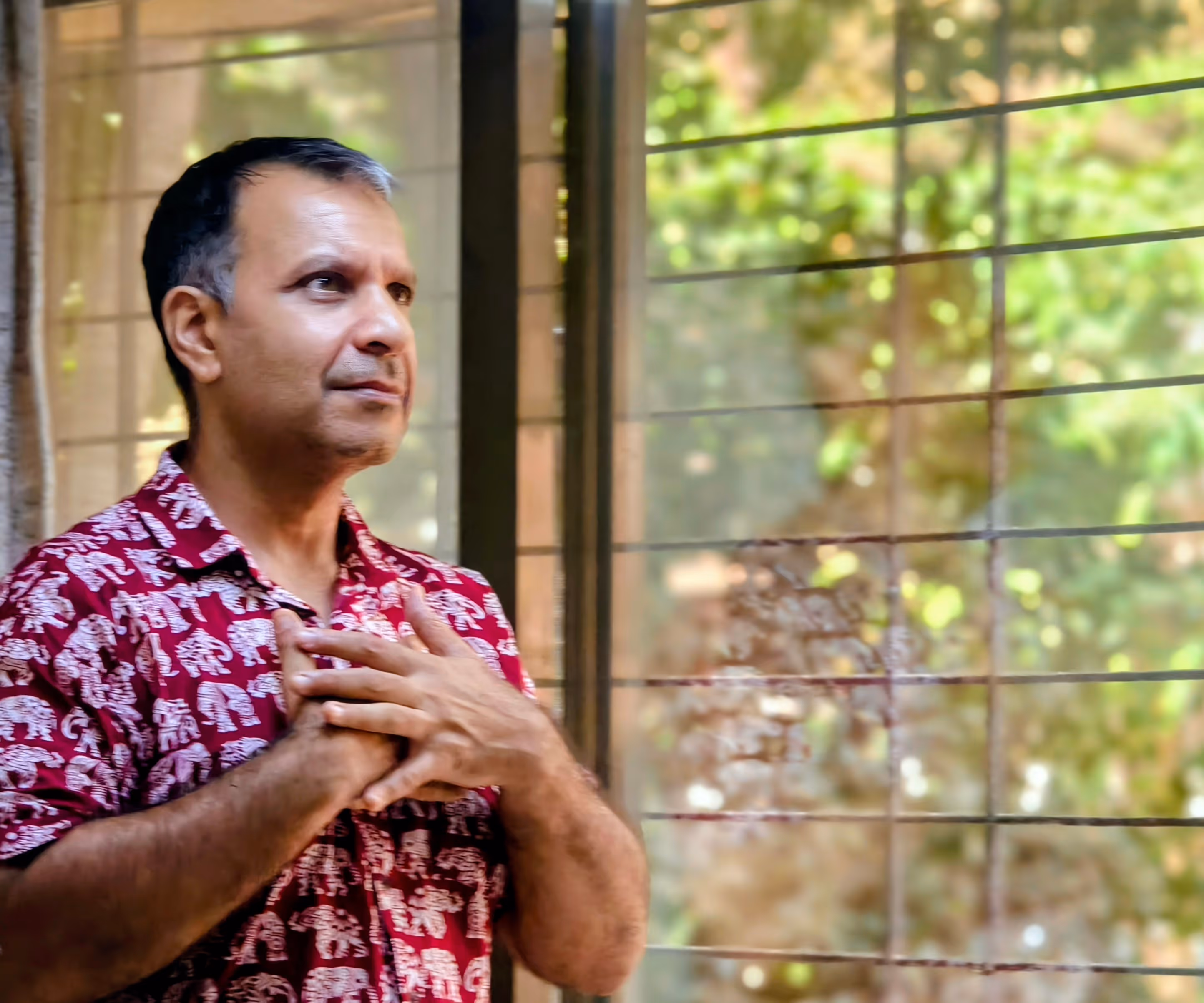 Man (Harsh Singh) in red elephant print shirt with hands on chest, standing next to window with blurred greenery outside, during Abhay Thhakkar's NLP course.