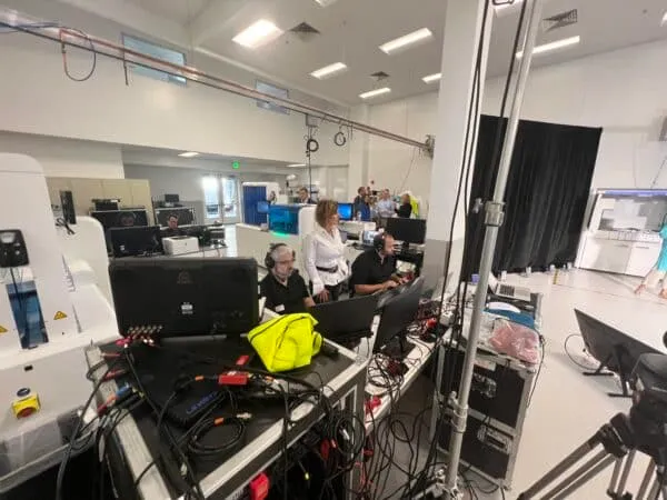 A livestreaming production crew working in a large room with multiple monitors, cables, and equipment, with a woman standing and two men seated wearing headsets.