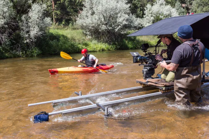 Person kayaking in shallow river while two cameramen film with a camera on a mounted rig in the water.