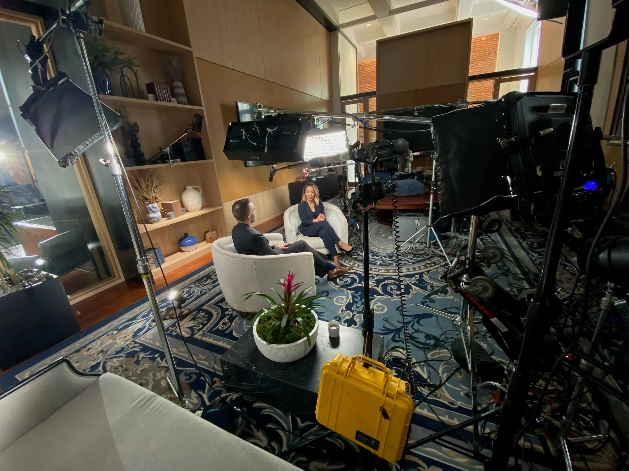 Two people seated in white armchairs facing each other in a well-lit room with film equipment and lights set up around them.