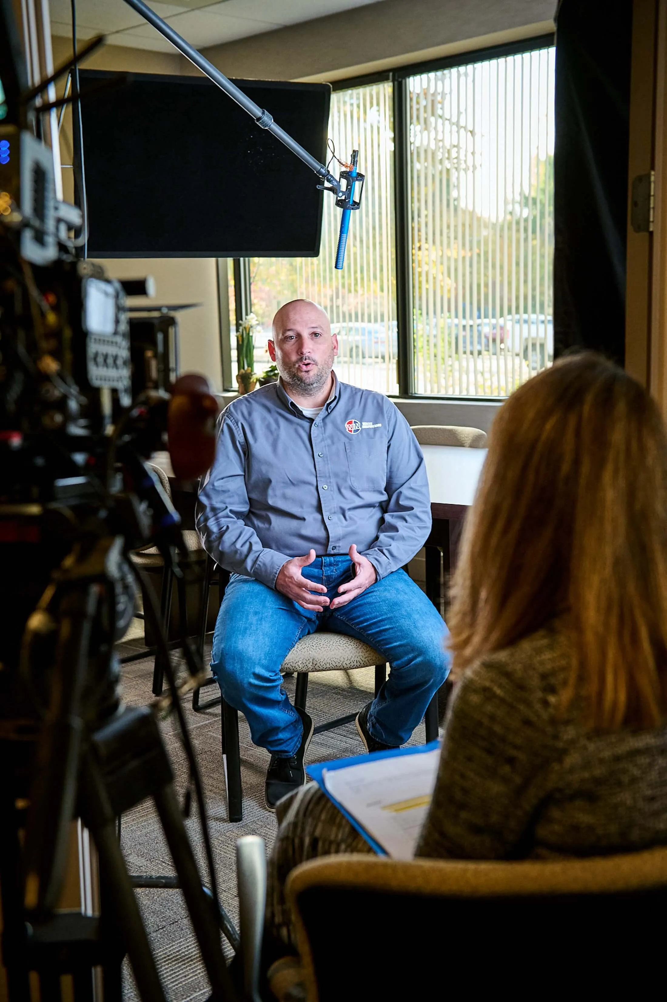 Man wearing a gray shirt and jeans is seated and speaking during a recorded interview in an office room with windows, facing a woman holding a clipboard.