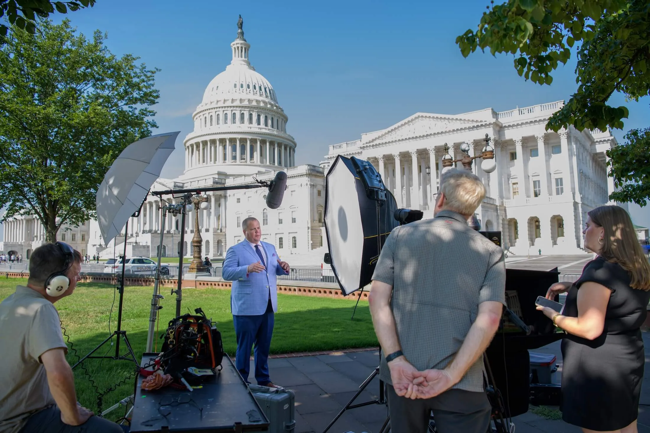 Interview setup near the U.S. Capitol with a man in a blue suit speaking on camera and crew operating lighting and sound equipment.