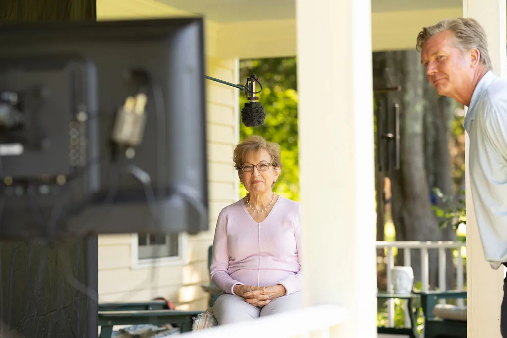 Elderly woman wearing glasses and a light pink sweater sitting on a porch, being filmed with a microphone overhead and a man observing nearby.