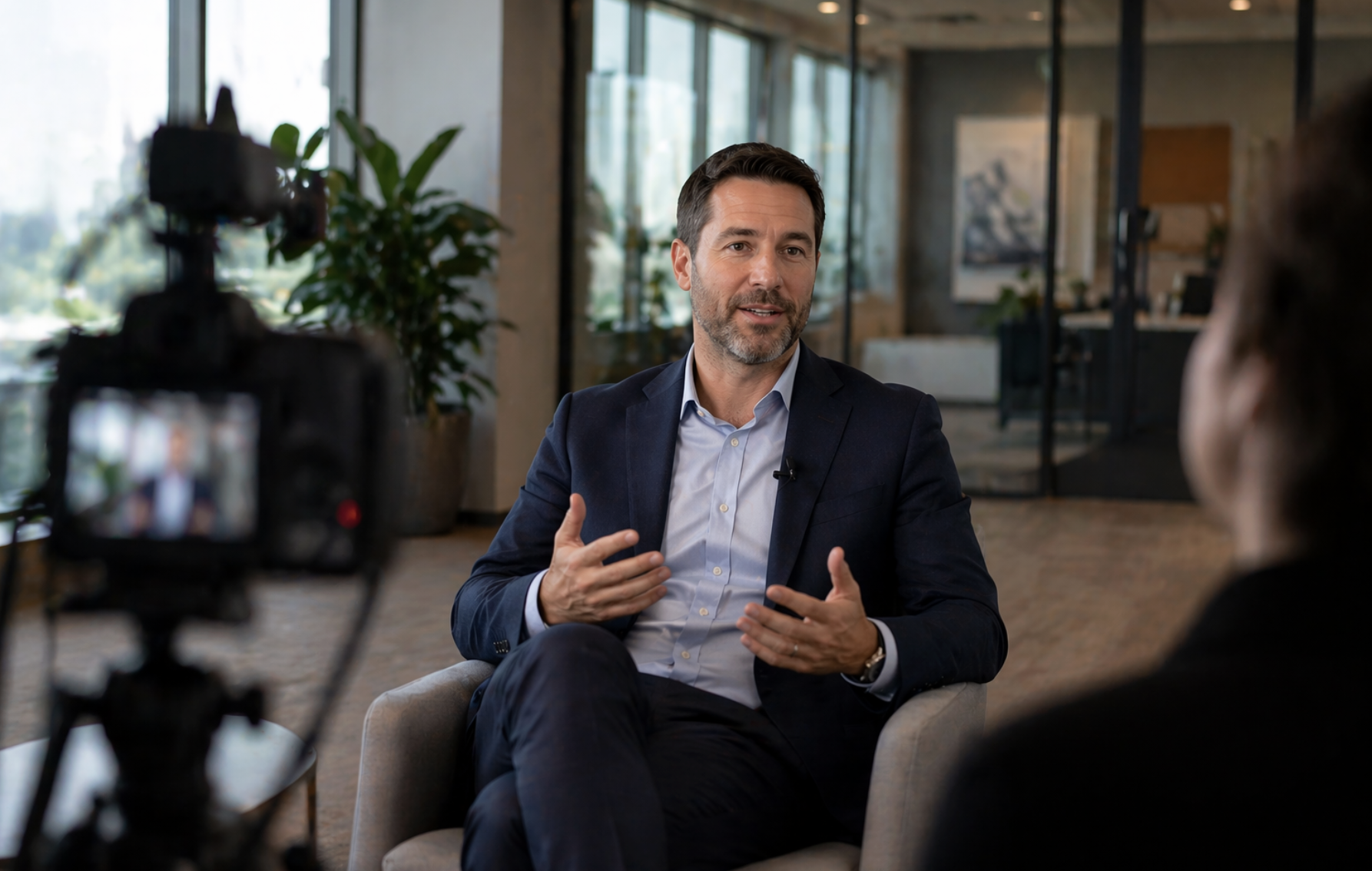 Man in a navy suit gesturing as he gives a testimonial interview, filmed by a camera in the foreground