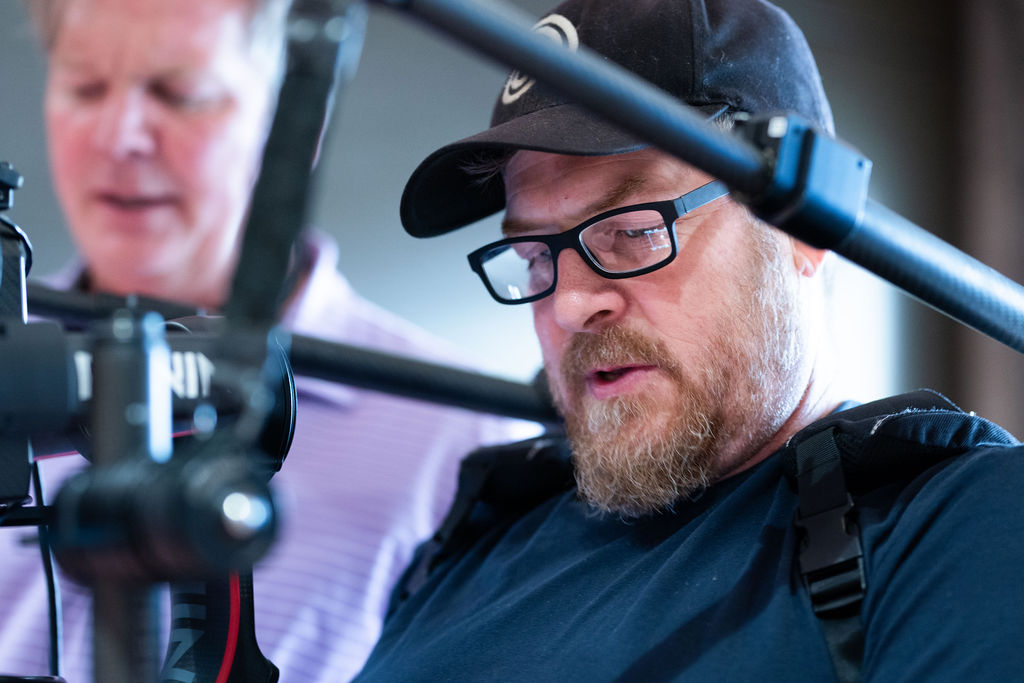 Close-up of a bearded camera operator in glasses and a cap working a camera rig