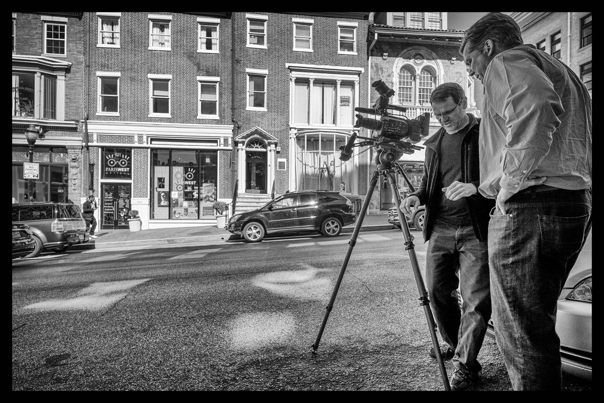 Black-and-white photo of a camera operator and colleague setting up a tripod-mounted camera on a city street