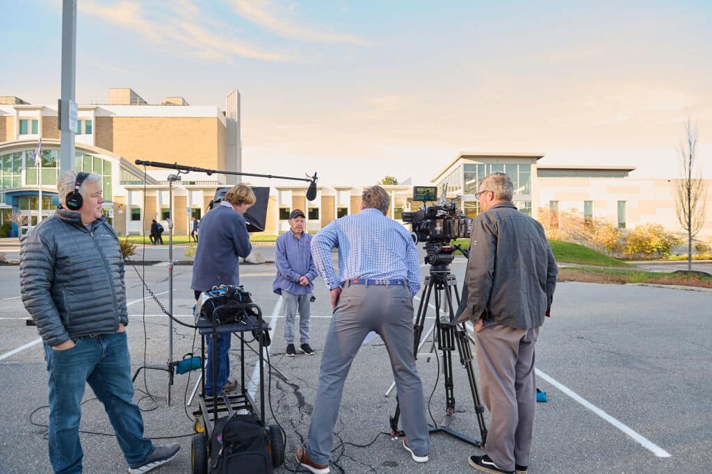Film crew with cameras, a boom mic, and headphones setting up a shot in a parking lot at sunset
