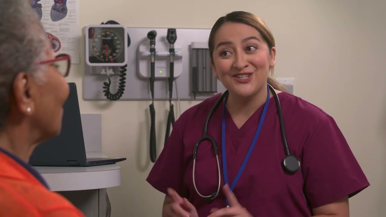 Smiling nurse in maroon scrubs with a stethoscope talking with an older patient in an exam room, from a CMS Medicare wellness visit eLearning module