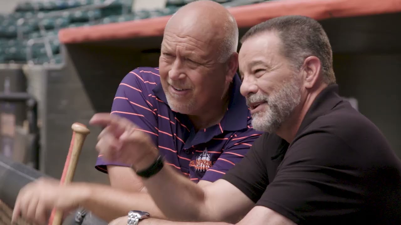 Two older men sharing a laugh in a baseball dugout, one holding a bat, from the Department of Labor ODEP Spanish-language PSA ¡Trabajar Funciona!