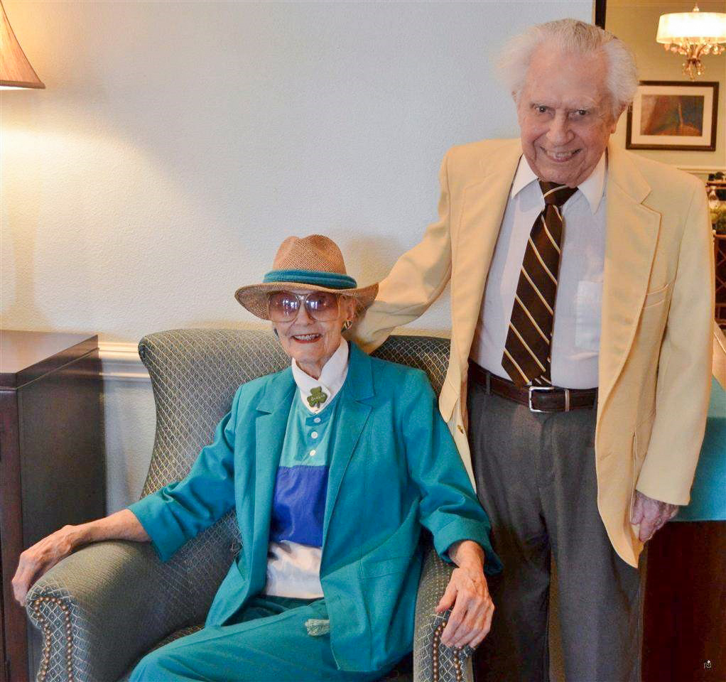 An elderly woman sitting in an armchair and an elderly man standing beside her, both smiling.