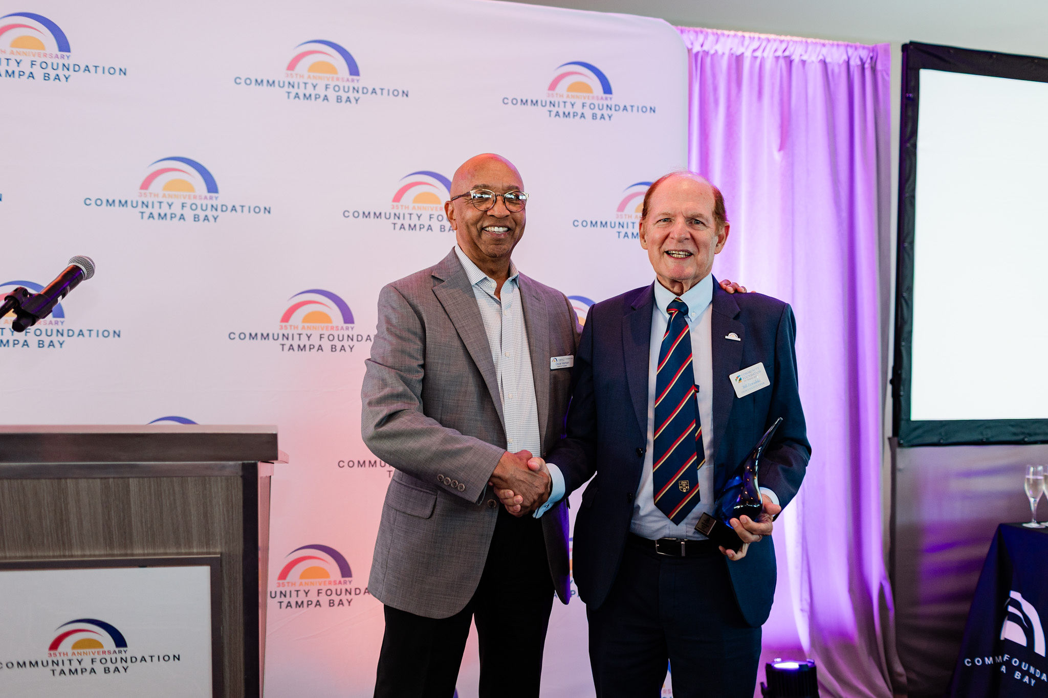 Two smiling men in suits shaking hands on a stage, with one holding an award.