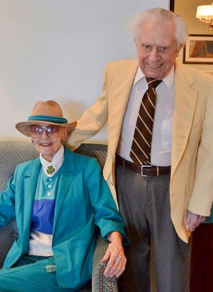 An elderly woman sitting in an armchair and an elderly man standing beside her, both smiling.