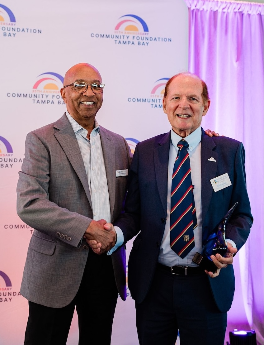 Two smiling men in suits shaking hands on a stage, with one holding an award.