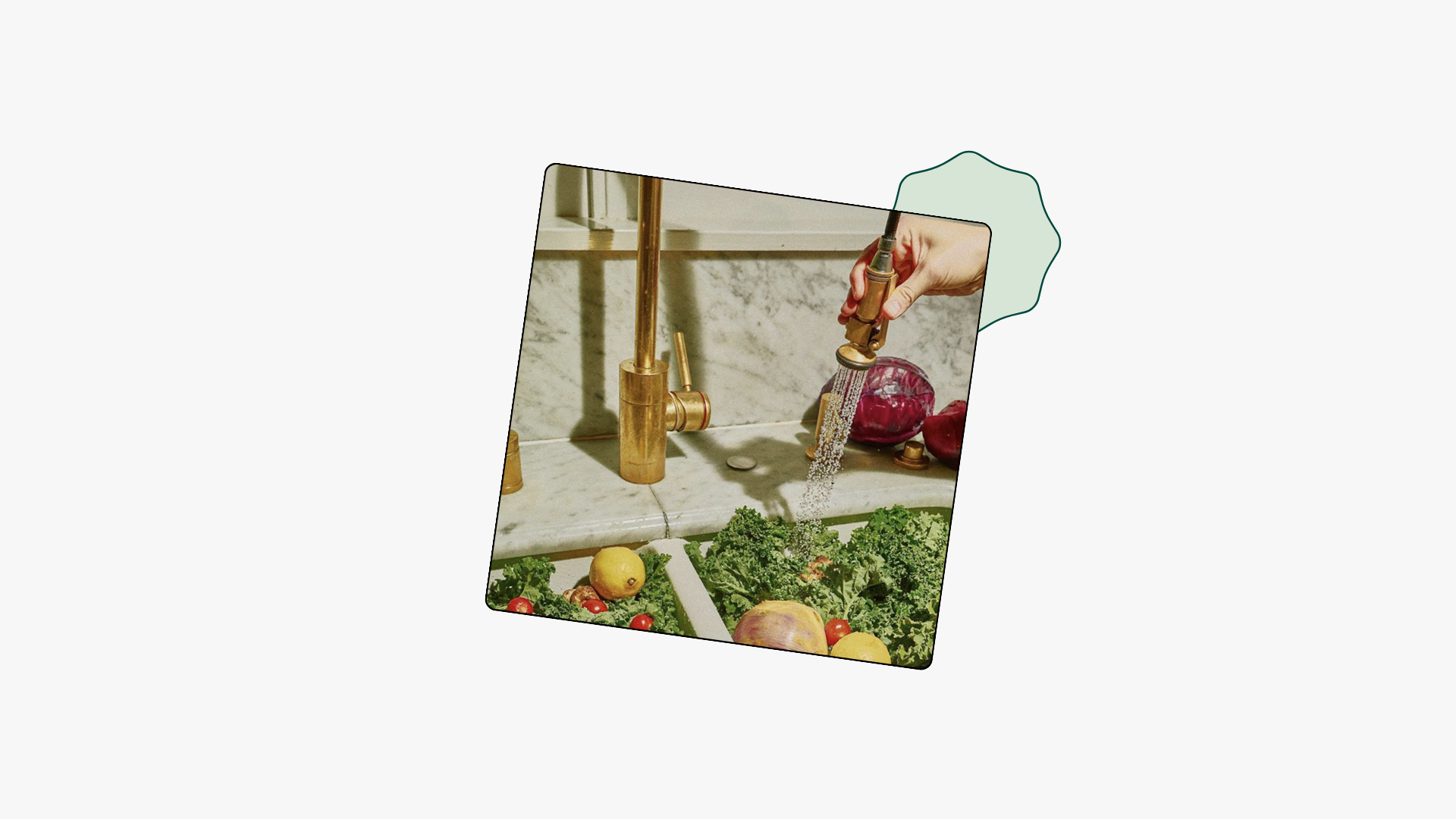 A hand uses a gold kitchen sprayer to rinse fresh vegetables including kale, tomatoes, and lemons in a marble sink.