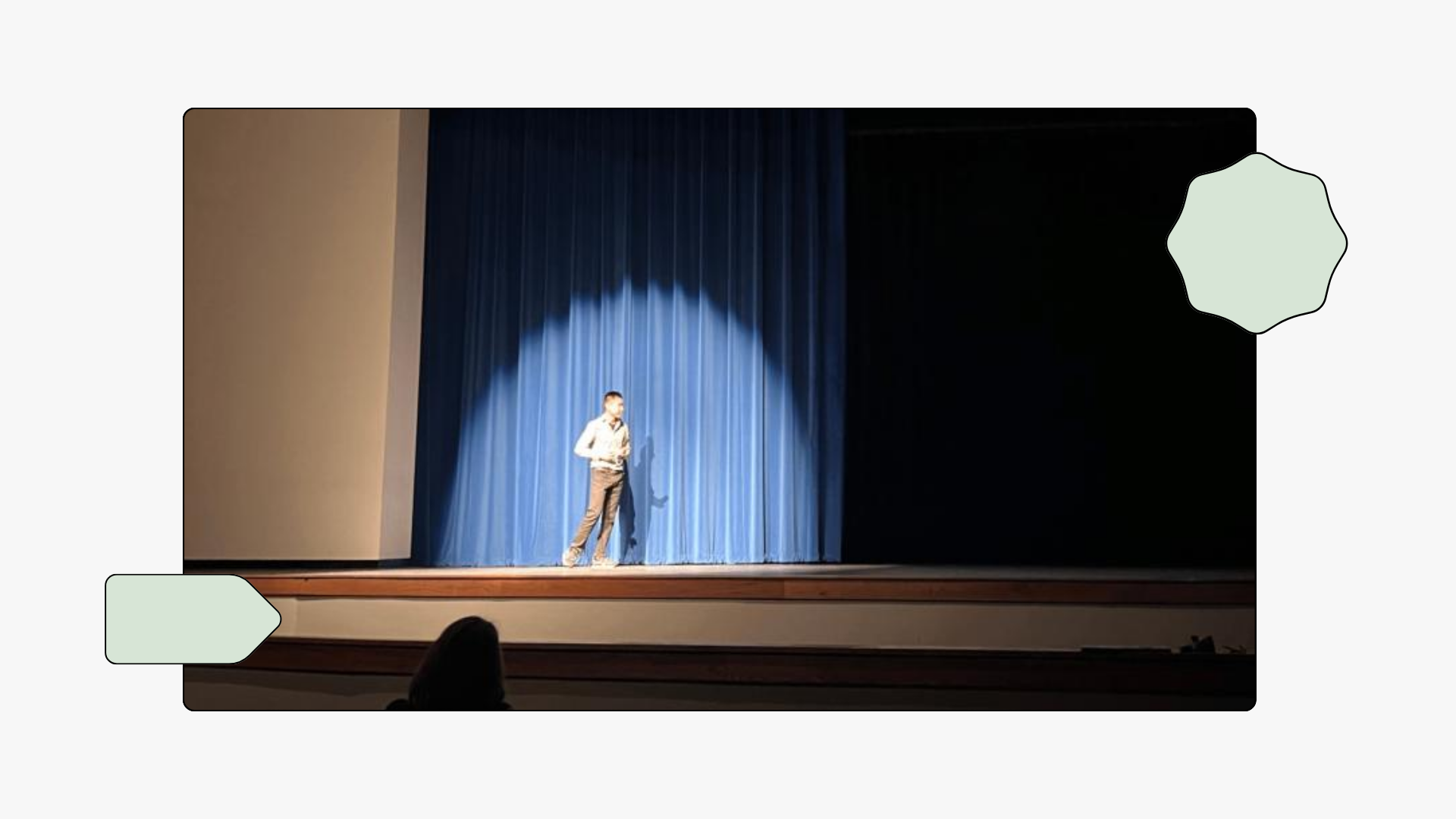 Joon stands alone on stage under a spotlight in front of closed blue curtains, presenting to an audience in a dimly lit auditorium.