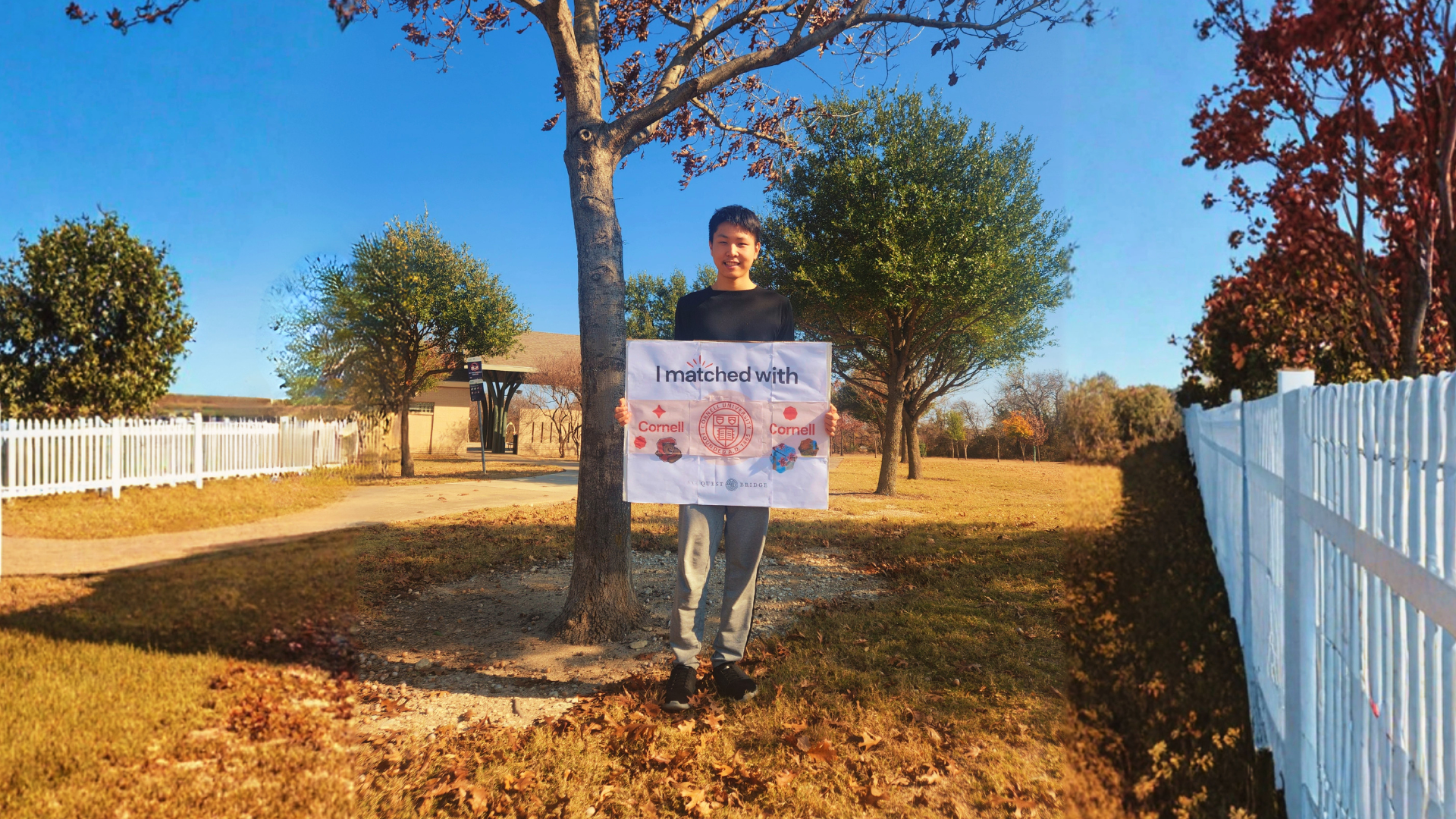 A wide shot of a student outdoors in a park holding a "I matched with Cornell" sign, surrounded by oak trees and autumn leaves on a sunny day.