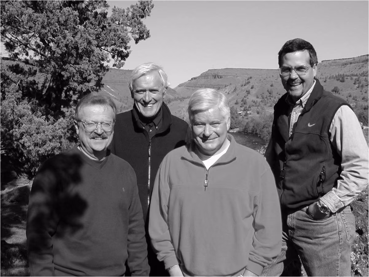 Four middle-aged men smiling outdoors with trees and hills in the background.