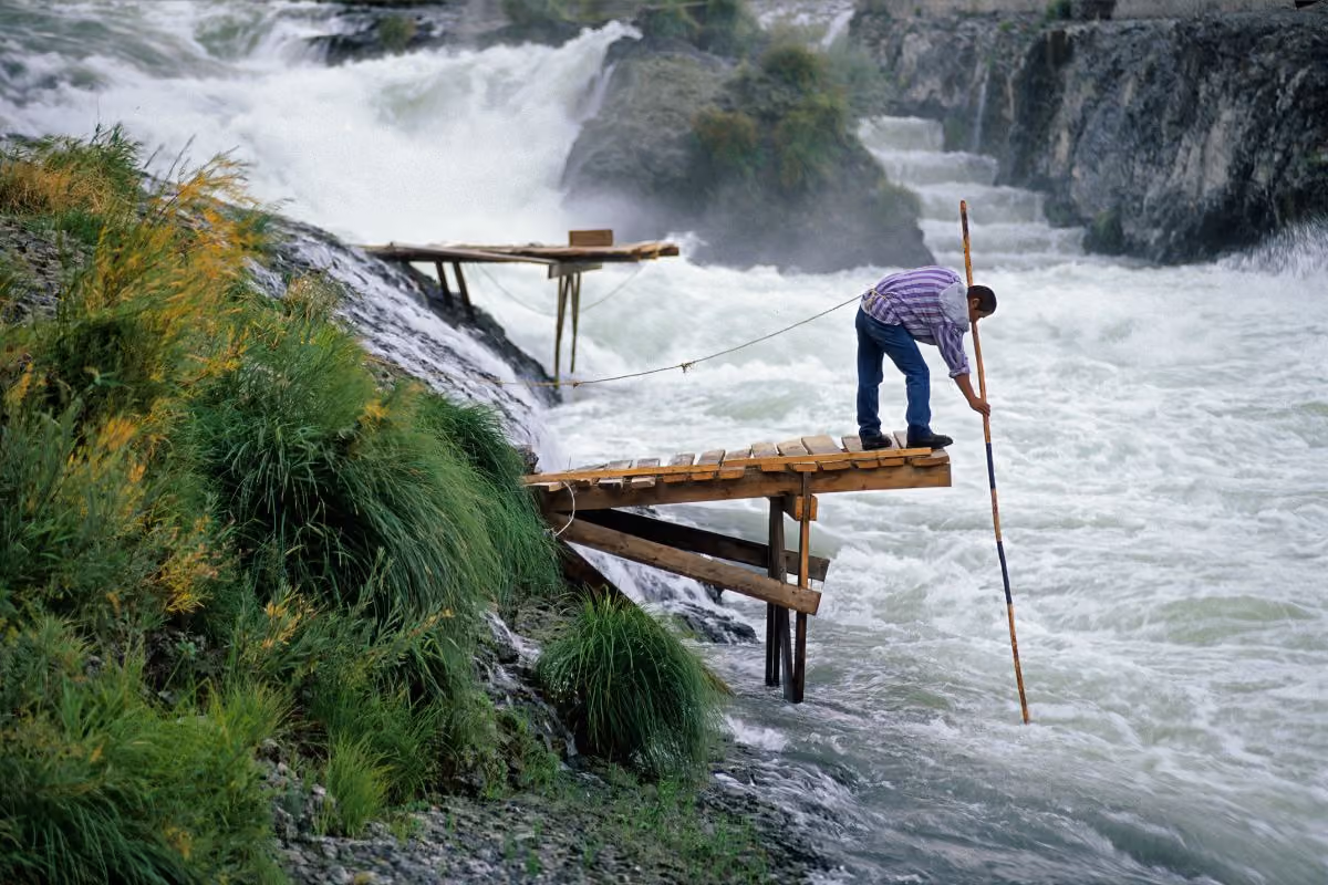 Man standing on wooden platform over turbulent river, holding a long pole and tethered by a rope.