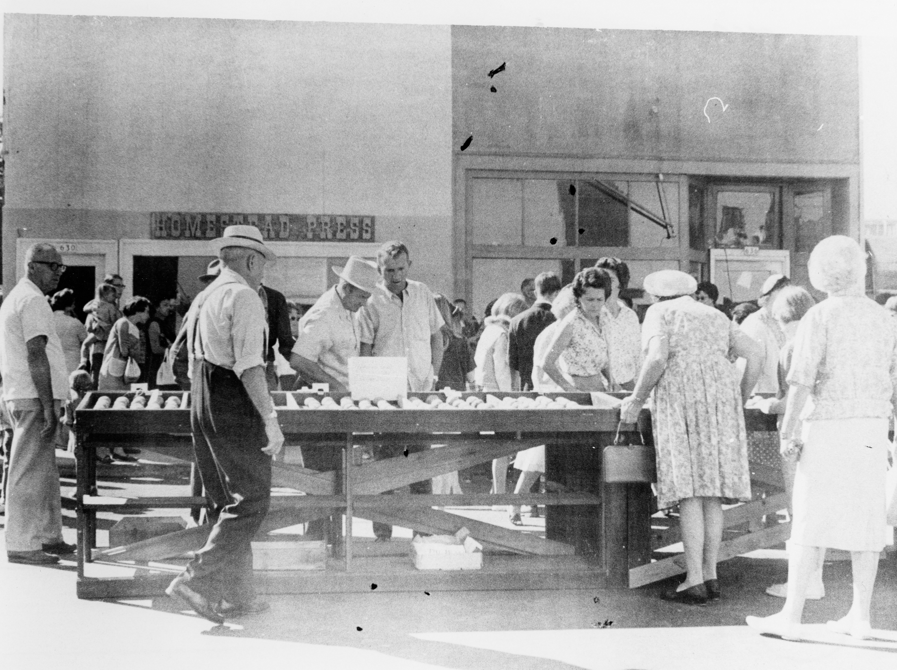An old black and white photograph of a crowd of well-dressed people looking through potatoes outside on a city street.