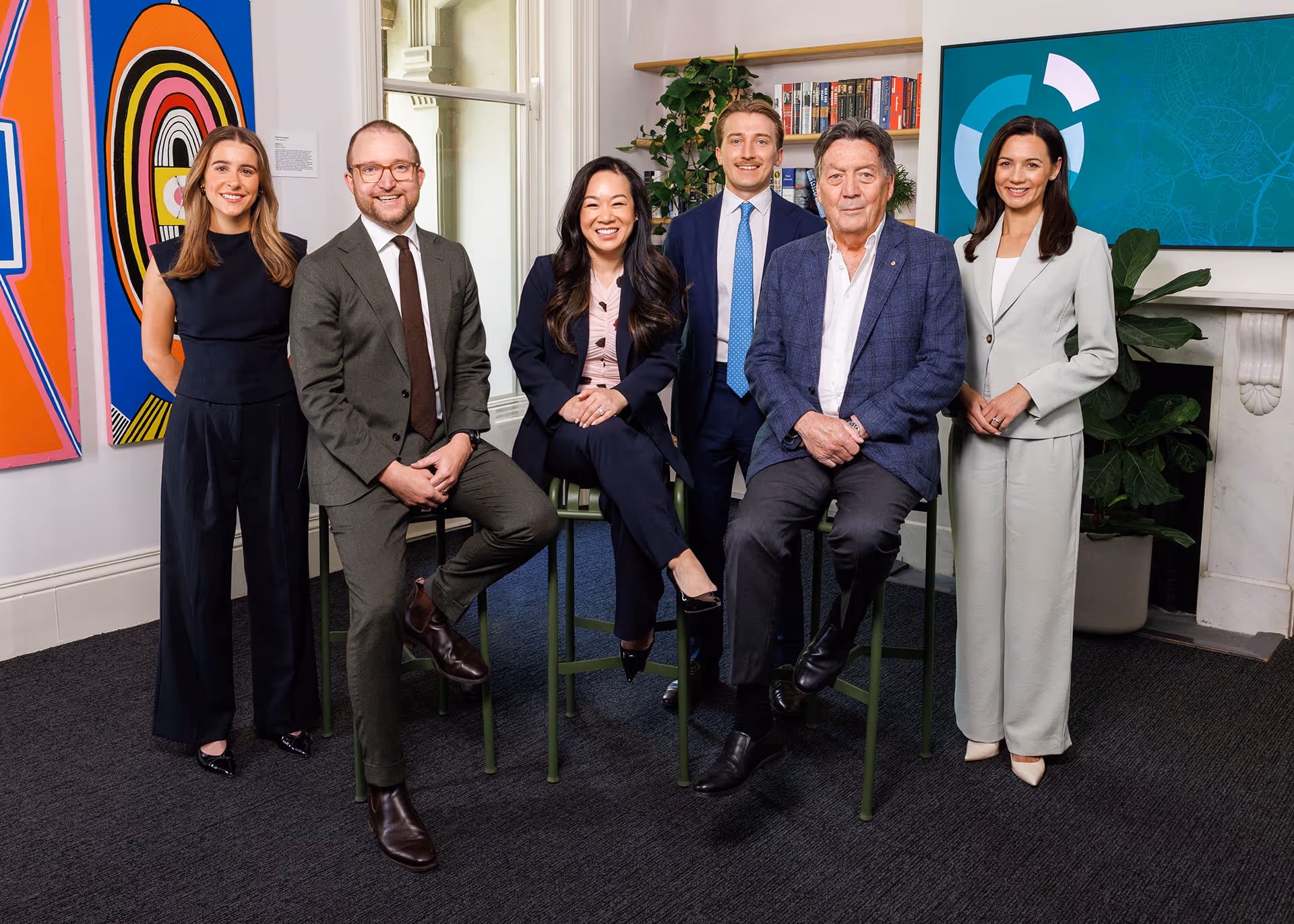 Six diverse professionals in business attire posing together in a modern office with colorful artwork and bookshelves.