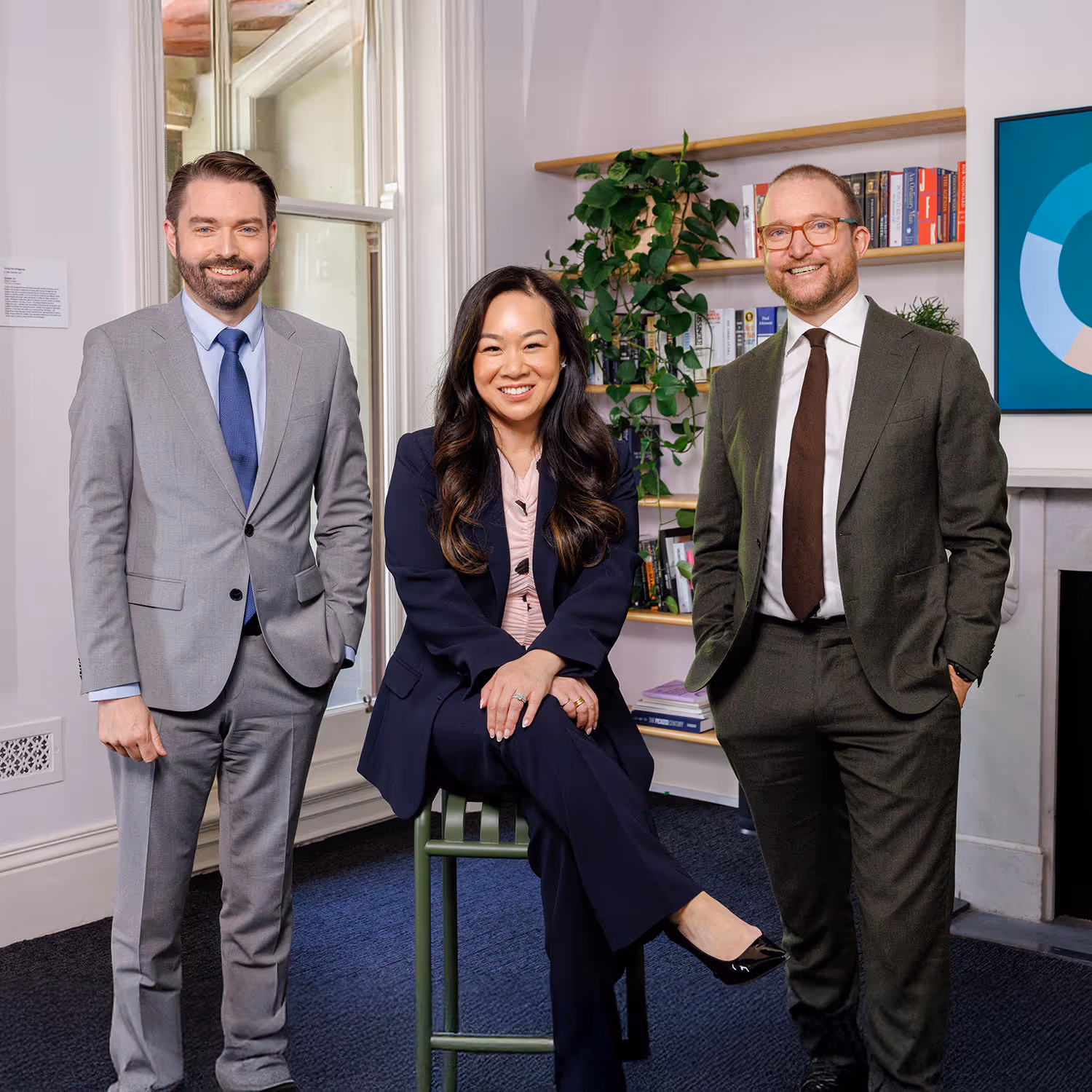 Three professionals in business attire smiling inside an office with bookshelves and a window.