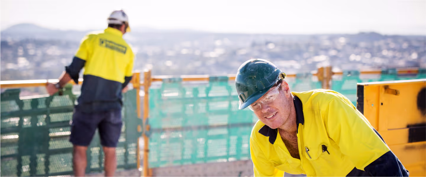 Two construction workers wearing yellow safety uniforms and helmets at a building site with a cityscape in the background.