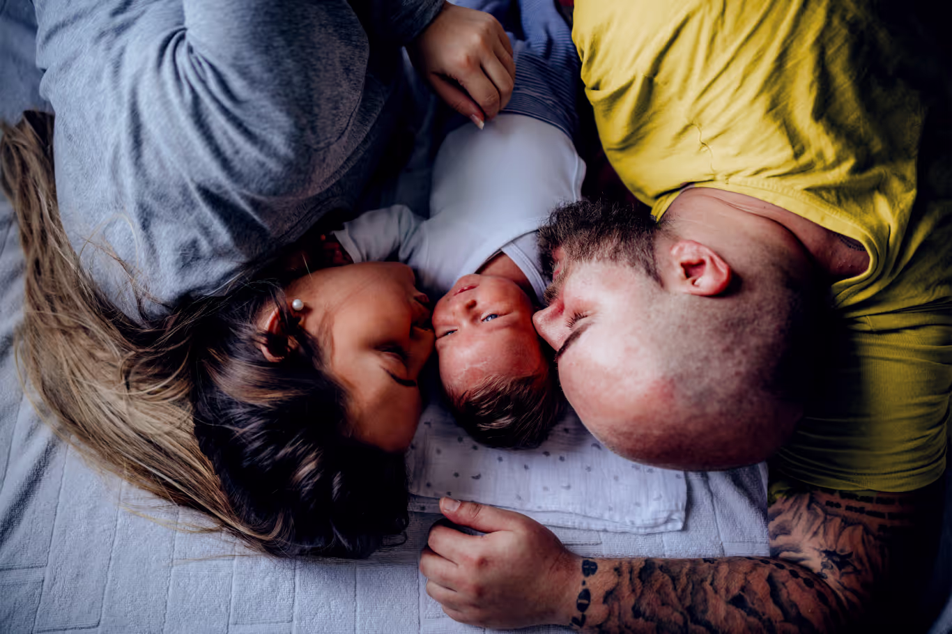 Parents lying on either side of their newborn baby on a bed, touching foreheads and looking lovingly at the baby.
