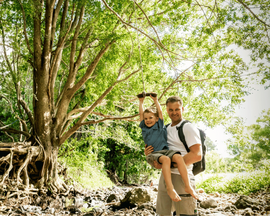 Man holding smiling child swinging on a rope swing in a sunny forest by a rocky stream.